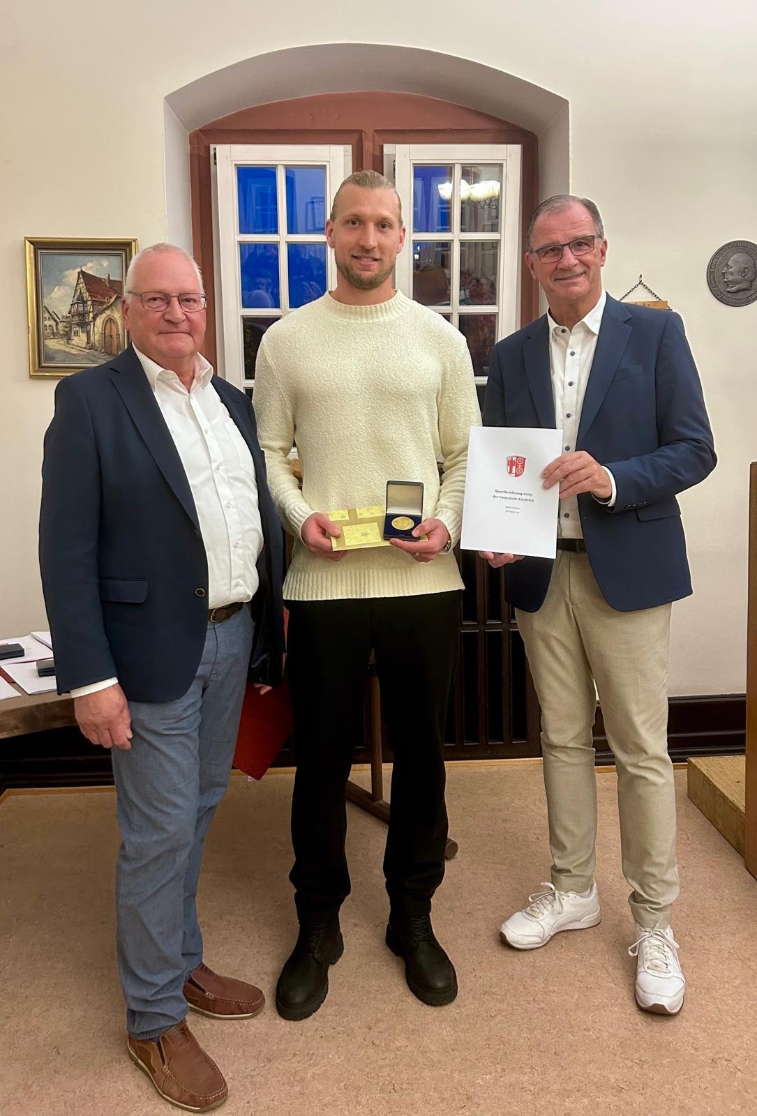 Drei Männer stehen vor einem weißen Sprossenfenster. Der Mann in der Mitte trägt einen beigen Pullover und eine dunkle Hose. Er trägt einen blonden Bart und hat eine goldene Urkunde und ein Geschenk in der Hand. Umrahmt wird er von zwei Männern mit Brillen,  weißen Hemden und blauen Jackets. Ein Mann hält eine weiße Urkunden-Mappe in der Hand. 