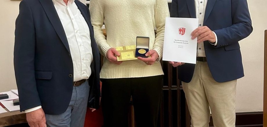 Drei Männer stehen vor einem weißen Sprossenfenster. Der Mann in der Mitte trägt einen beigen Pullover und eine dunkle Hose. Er trägt einen blonden Bart und hat eine goldene Urkunde und ein Geschenk in der Hand. Umrahmt wird er von zwei Männern mit Brillen,  weißen Hemden und blauen Jackets. Ein Mann hält eine weiße Urkunden-Mappe in der Hand. 
