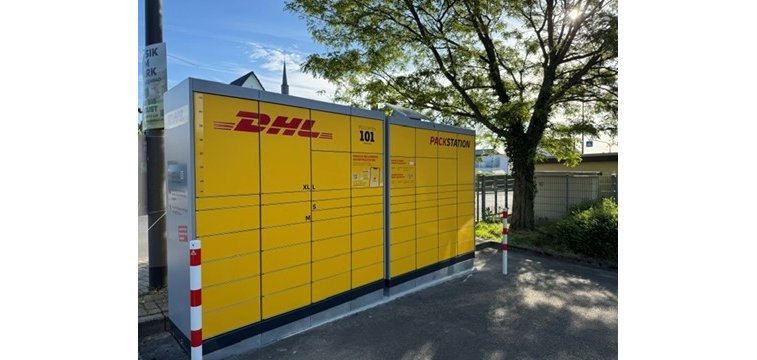 Man sieht eine gelbe Packstation mit geschlossenen Fächsern und der Schrift in rot und schwarz. Im Hintergrund ist ein Baum und blauer Himmel. Im Vordergrund zwei Pfosten links und rechts von der Packstation in weiß-rot und Asphaltboden.