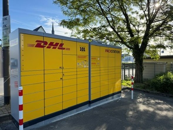 Man sieht eine gelbe Packstation mit geschlossenen Fächsern und der Schrift in rot und schwarz. Im Hintergrund ist ein Baum und blauer Himmel. Im Vordergrund zwei Pfosten links und rechts von der Packstation in weiß-rot und Asphaltboden.