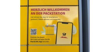 Man sieht ein gelbes Fach einer Packstation, af dem ein QR-Code und ein Handy mit gelbem Display und einem schwarzem Posthorn sowie dem rotem DHL-Logo. Im HIntergrund ist eine gelbe Packstation mit gezeichneten Büschen, Bäumen und Häusern.