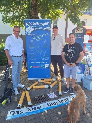 Drei Männer stehen neben einem Roll-Up in blau. Davor liegen überdimensionale Zigarettenkippen und ein Banner in weiß und blau mit der Aufschrift "Das Meer beginnt hier." Ein Mann hält einen braunen Hund an einer Leine. Der Hund steht mit dem Rücken zum BIld.