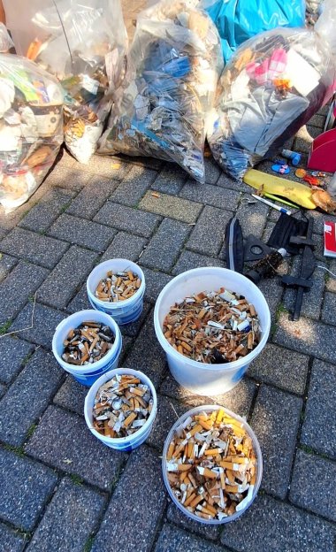 Auf einer geplasterten Fläche stehen fünf weiße Eimer, die mit Zigarettenkippen gefüllt sind. Dahinter liegen mehrere Müllsäcke, die mit Müll gefüllt sind.
