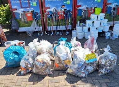 Vor vier Altkleidercontainern stehen weiße Eimer zu einer Pyramide gestapelt und mehrere Greifer zum Müllsammeln. Im Vordergrund liegen mehrere gefüllte Müllsäcke.