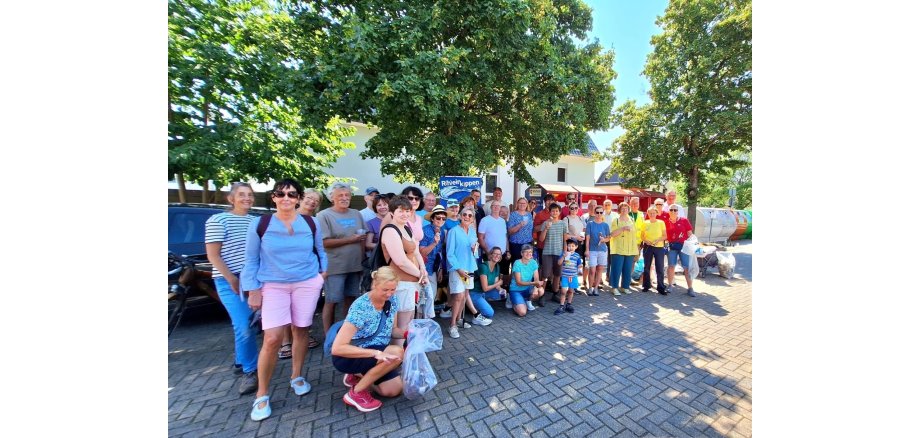 Mehrere Personen stehen und knieen auf einer gepflasteren Fläche. IM Hintergrnd sind belaubte Bäume.  