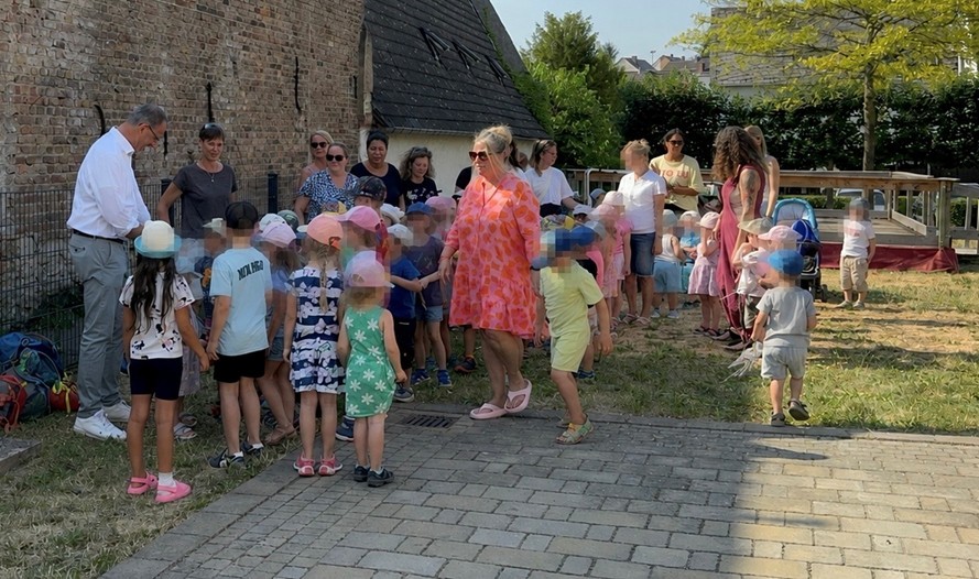 Auf einer Wiese und einer gepflasterten Fläche, stehen mehrere Kinder und Frauen bei einem Mann mit weißem Hemd und grauer Hose. Die Kinder tragen Kappen gegen die Sonne.