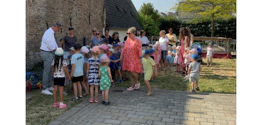 Auf einer Wiese und einer gepflasterten Fläche, stehen mehrere Kinder und Frauen bei einem Mann mit weißem Hemd und grauer Hose. Die Kinder tragen Kappen gegen die Sonne.
