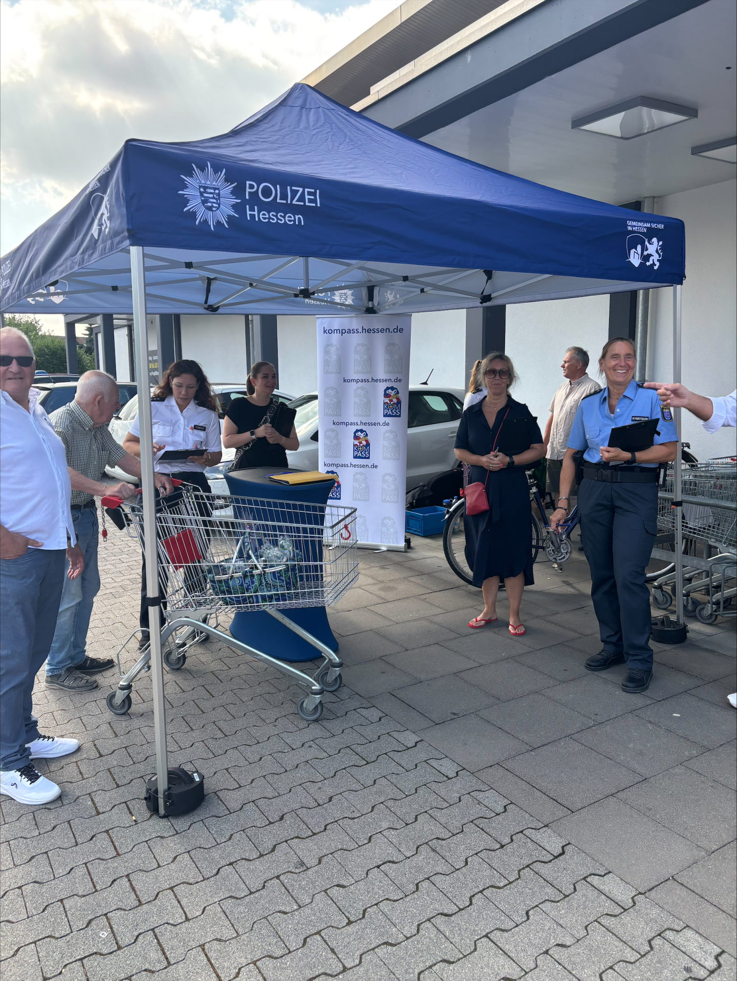 Unter einem blauen Pavillon der Polizei Hessen steht ein Mann mit Einkaufswagen, zwei Polizistinnen, zwei Frauen in schwarz gekleidet und ein Herr mit weißem Hemd und blauer Jeans. Eine Leinwand ist ausgerollt und ein Stehtisch mit dunkler Husse steht unter dem Pavillon.