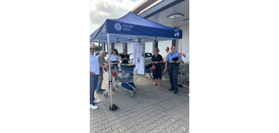 Unter einem blauen Pavillon der Polizei Hessen steht ein Mann mit Einkaufswagen, zwei Polizistinnen, zwei Frauen in schwarz gekleidet und ein Herr mit weißem Hemd und blauer Jeans. Eine Leinwand ist ausgerollt und ein Stehtisch mit dunkler Husse steht unter dem Pavillon.
