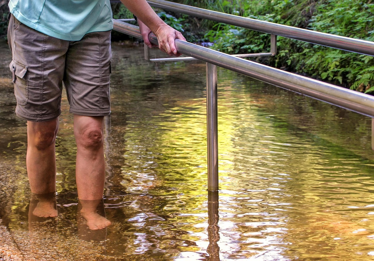 In einem gefülltem Wasserbecken, steht ein Mann und hält sich an einer Metallstange fest. Er trägt eine kurze graue Hose mit Seitentaschen und ein hellblaues Shirt. Man sieht keinen Oberkörper.