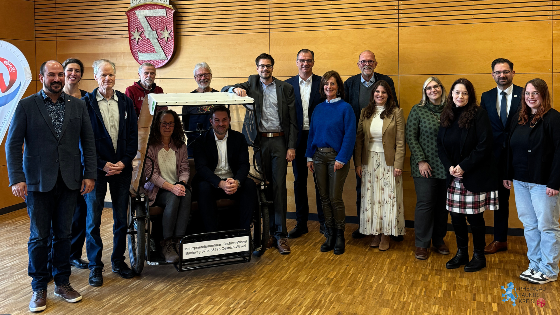 Vor einer mit Holz verkleideten Wand stehen 8 Männer und 6 Frauen im Halbkreis. Im Halbkreis ist eine Fahrradrikscha für zwei Personen mit beigem Dach, in der eine Frau und ein Mann sitzen, integriert. An der Wand hängt ein rotes Wappen mit einem spiegelverkehrtem "Z" und jeweils einem Stern links und rechts davon. Der Boden ist ein Parkettboden mit kleinen Rechtecken.