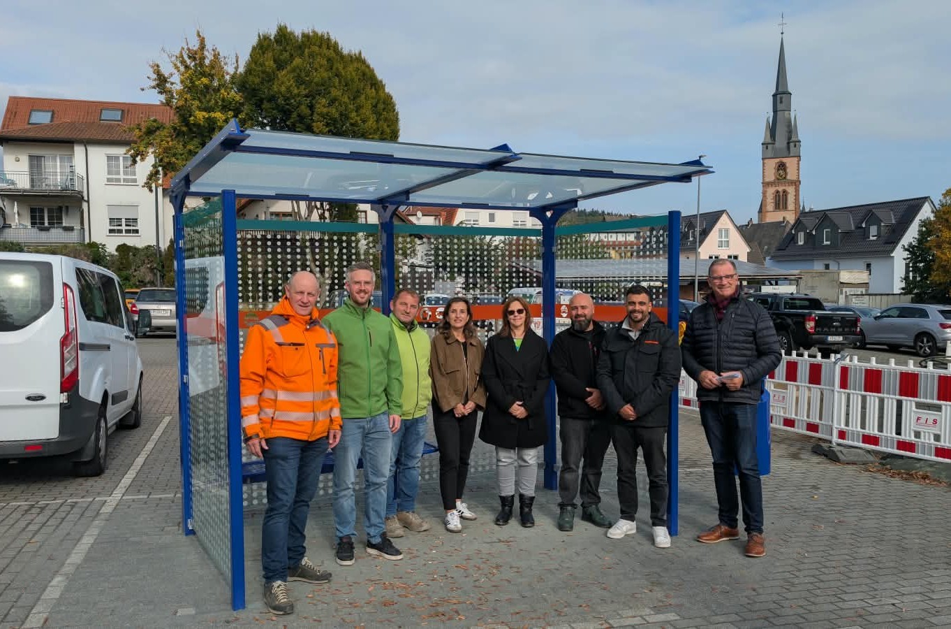 Vor einer Bushaltestelle mit blauem Metallgestänge und Glasflächen, stehen sechs Männer und zwei Frauen im Halbkreis. Im Hintergrund parken Autos und der Kirchturm ist zu sehen.