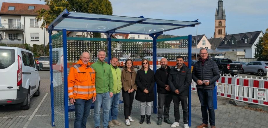 Vor einer Bushaltestelle mit blauem Metallgestänge und Glasflächen, stehen sechs Männer und zwei Frauen im Halbkreis. Im Hintergrund parken Autos und der Kirchturm ist zu sehen.