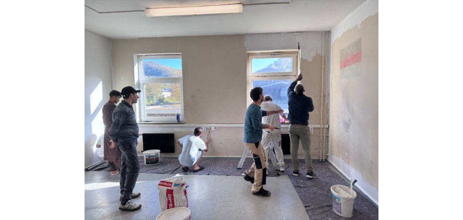 In einem Raum mit zwei Fenstern brennt eine rechteckige Lampe an der Decke und der Boden ist am Rand mit Malerflies ausgelegt. An einer Wand ist eine Deutschlandfahne aufgemalt. Man sieht sieben Männer, die zusammen den Raum mit weißer Farbe streichen.