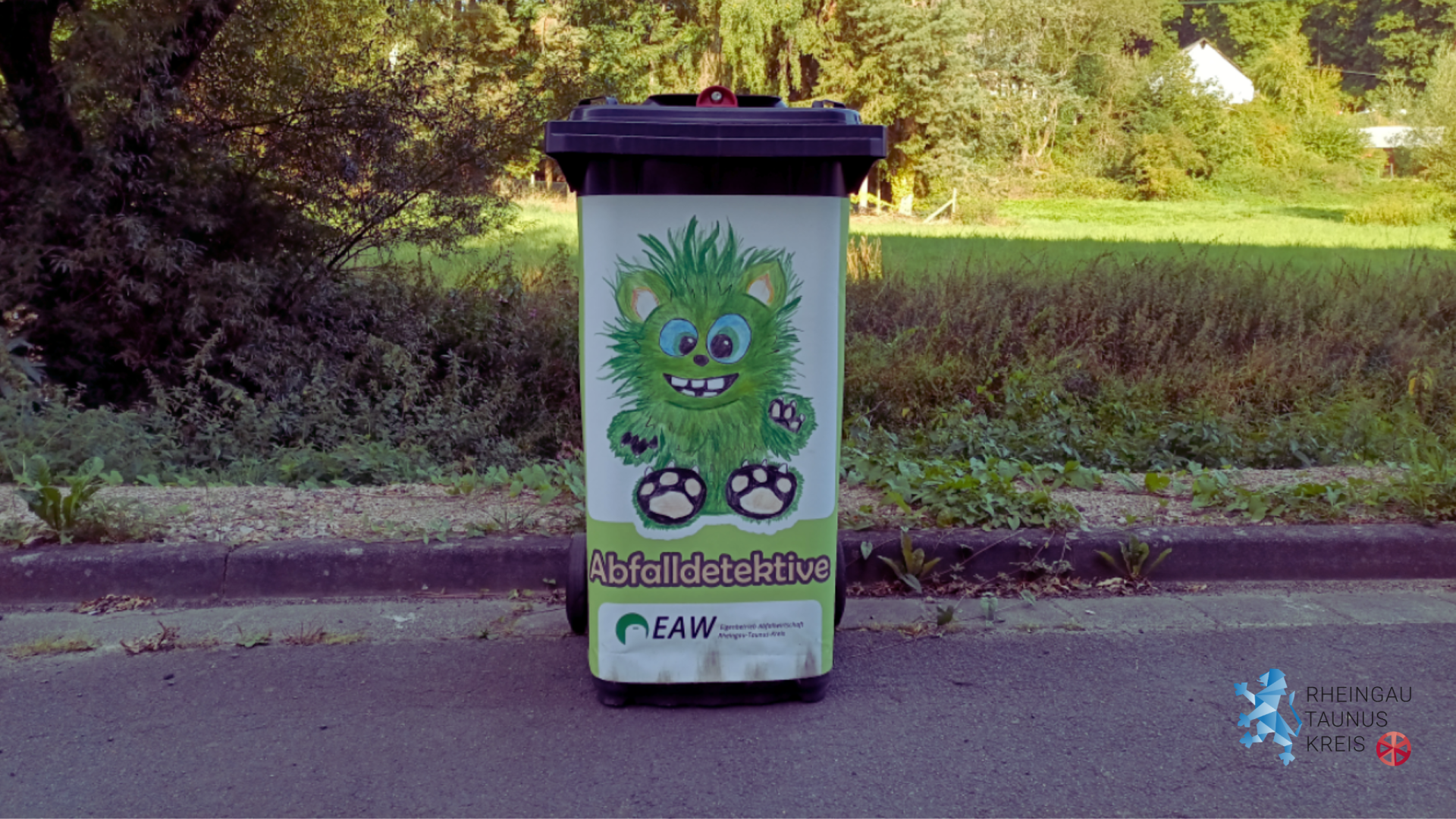 Auf einer Straße steht eine schwarze Tonne, auf dem ein Plakat klebt, auf dem vor einem weißem Hintergrund ein gezeichnetes, Monster mit großen blauen Augen, grünem Fell, großen Zähnen und Tatzen gezeichnet ist. Darunter steht Abfalldetektive und das Logo von EAW. Im HIntergrund der Tonne sind Unkraut, Hecken, Wiesen und Bäume zu sehen.