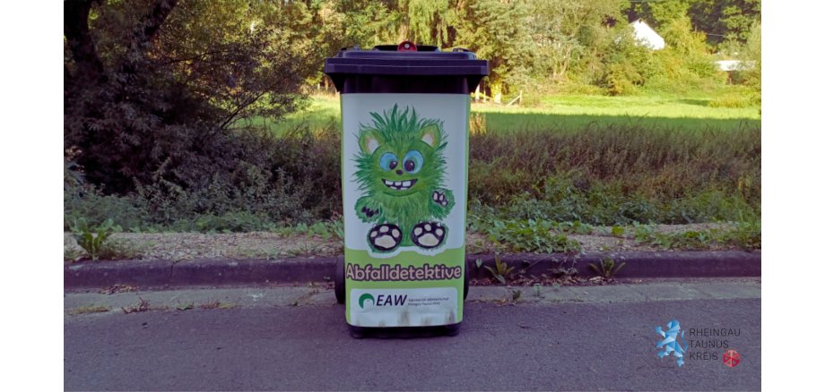 Auf einer Straße steht eine schwarze Tonne, auf dem ein Plakat klebt, auf dem vor einem weißem Hintergrund ein gezeichnetes, Monster mit großen blauen Augen, grünem Fell, großen Zähnen und Tatzen gezeichnet ist. Darunter steht Abfalldetektive und das Logo von EAW. Im HIntergrund der Tonne sind Unkraut, Hecken, Wiesen und Bäume zu sehen.