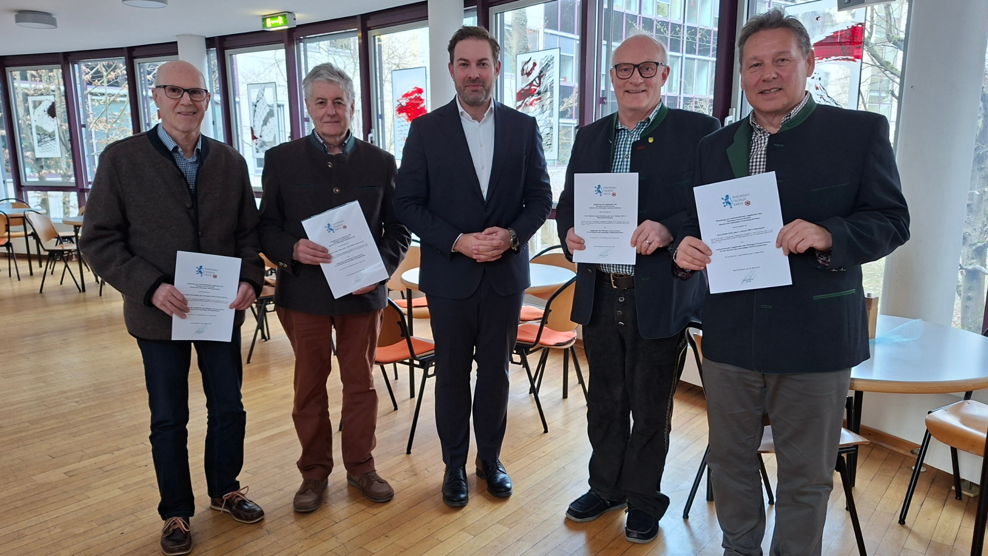 Kommunalwahl - 9 Vor einer Fensterfront und mehreren runden Tischen mit Stühlen, stehen fünf Männer in dunkler Kleidung und die jeweils zwei äußeren Personen halten weiße Urkunden in den Händen.