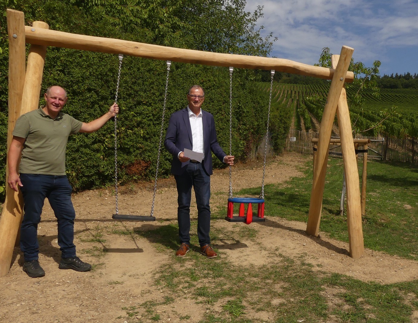 An einem breiten Gerüst aus Holzstämmen hängen an silbernen Ketten eine Schaukel und eine Babyschaukel. Zwei Männer halten jeweils eine Kette fest. Im Hintergrund sind Büsche, ein Holzzaun und Weinberge zu erkennen.