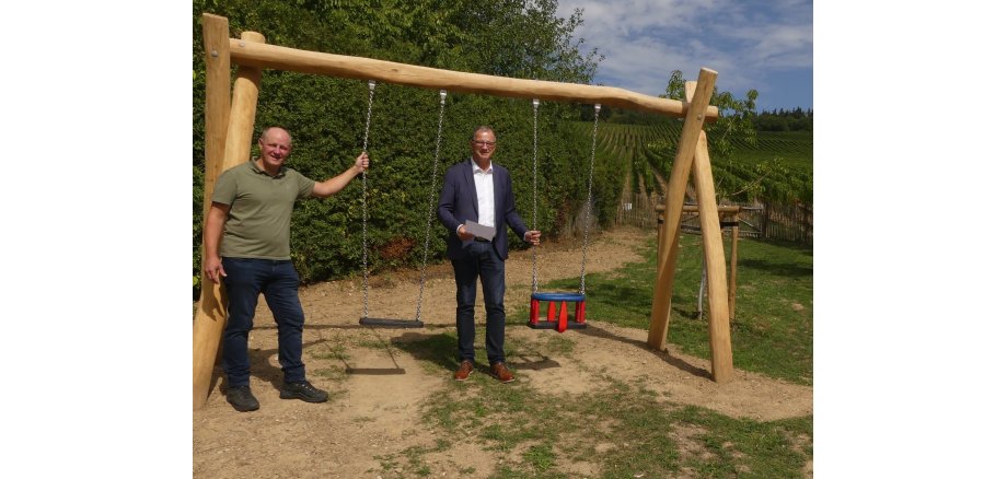 An einem breiten Gerüst aus Holzstämmen hängen an silbernen Ketten eine Schaukel und eine Babyschaukel. Zwei Männer halten jeweils eine Kette fest. Im Hintergrund sind Büsche, ein Holzzaun und Weinberge zu erkennen.