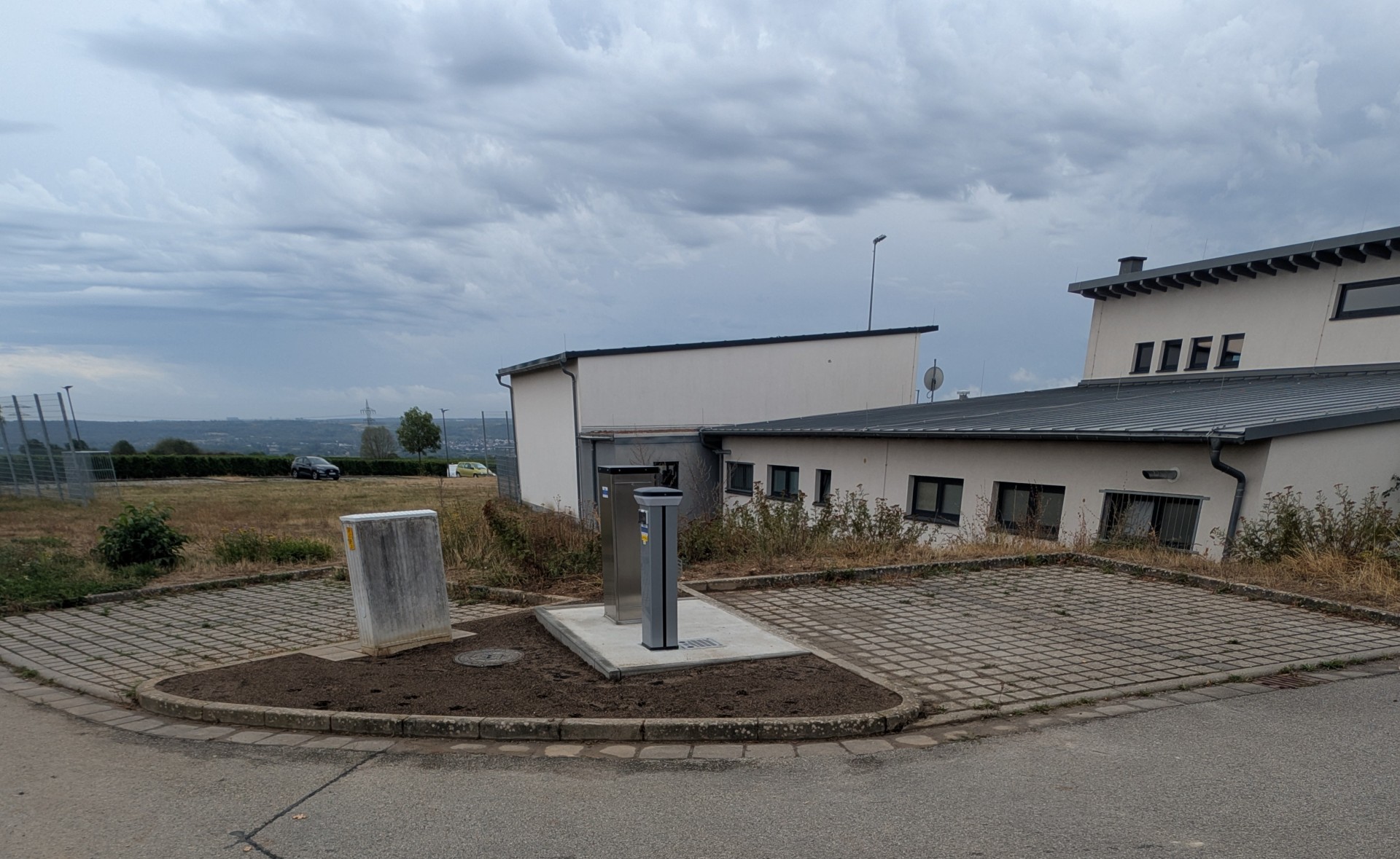 Je zwei gepflasterte Parkplätze, im rechten Winkel und dazwischen auf einem dreieckigem Erdstück steht ein Stromkasten und zwei silberne Säulen. Im Hintergrund sind beige Gebäude mit Fenstern.