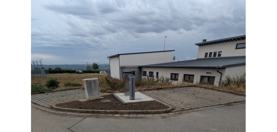 Je zwei gepflasterte Parkplätze, im rechten Winkel und dazwischen auf einem dreieckigem Erdstück steht ein Stromkasten und zwei silberne Säulen. Im Hintergrund sind beige Gebäude mit Fenstern.