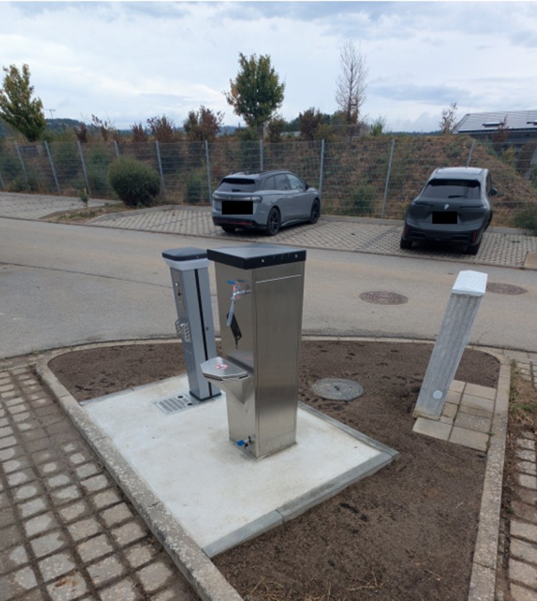 Auf einem dreieckigem Erdstück steht ein Stromkasten und zwei silberne Säulen. Daneben sind gepflastere Parkplätze. Gegenüber sind mehrere Parkplätze, auf denen zwei Autos parken. Dahinter ist ein langer Erdhügel, der mit Bäumen und Strächern bepflanzt ist und ein Hausdach ist erkennbar.