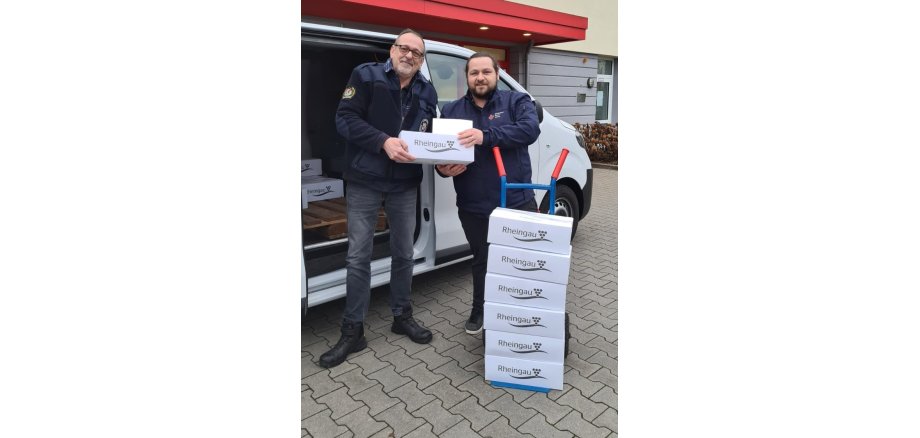 Vor einem weißen Lieferwagen mit geöffneter Schiebetür stehen zwei Männer mit Winterjacken und überreichen sich eine Kiste Wein. Im Lieferwagen liegt eine Palette mit weiteren weißen Weinkisten. Vor dem Lieferwagen steht eine Sackkarre mit sechs weißen Kisten Wein.