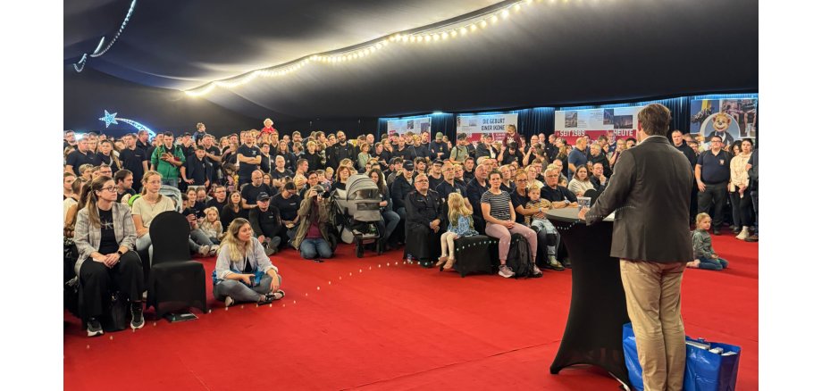 Newsfeed Bild - Dankesfest In einem großen Zelt mit schwarzen Decken und Wänden, sitzen und stehen viele Personen auf Stühlen. Der Boden ist mit einem rotem Teppich bedeckt. Vorn steht ein Mann an einem Bistrotisch mit einer schwarzen Husse.