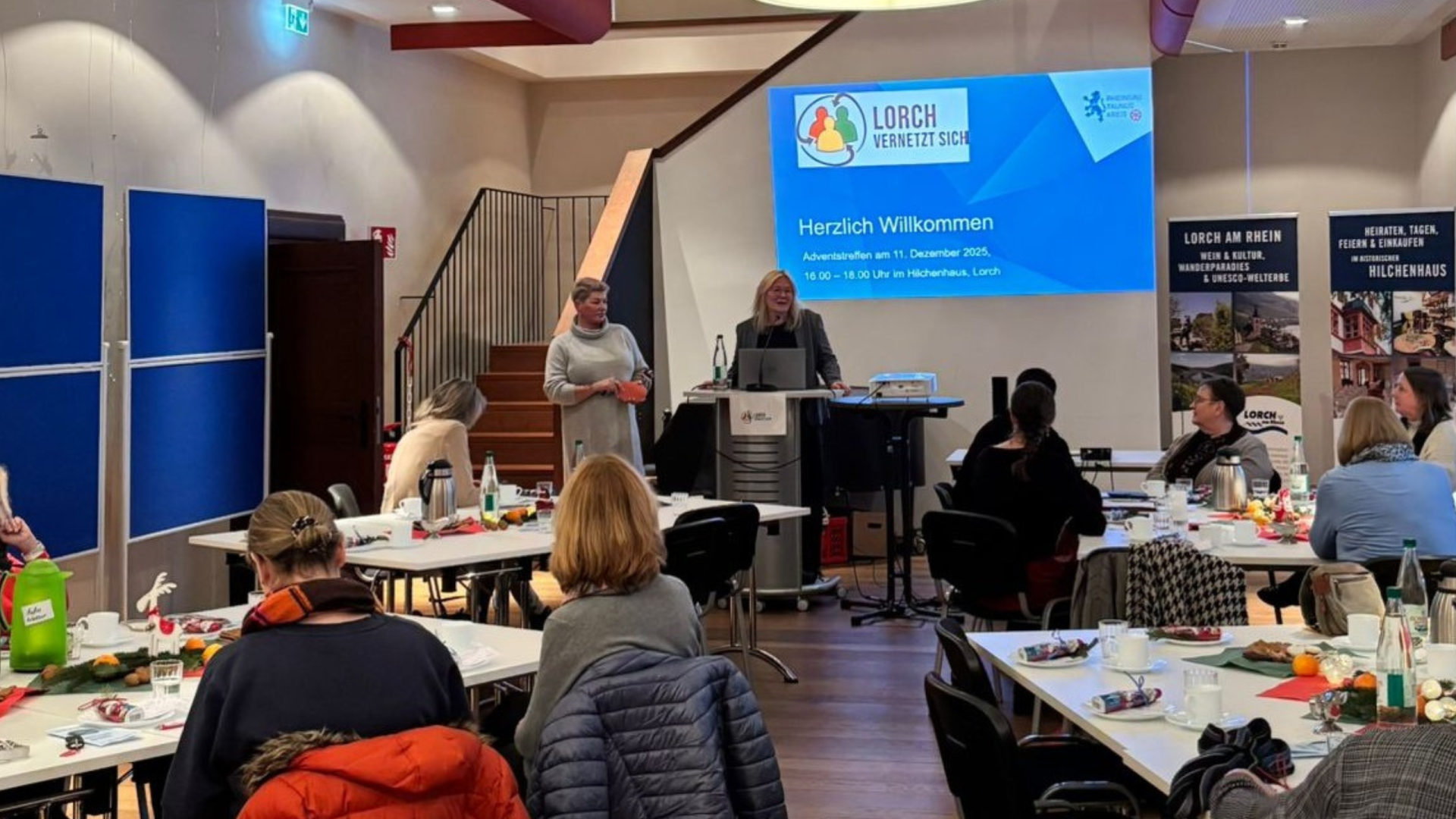 An vier rechtekigen Tischen, die mit Servietten, Mandarinen, Nüssen und Tannenzweigen dekoriert sind, sitzen mehrere Frauen. Vorn ist ein Rednerpult und ein Beamer auf einem Bistrotisch. Vorn stehen zwei Frauen am und neben dem Rednerpult. Hinter ihnen an der Wand ist das Titelbild einer Präsentation. Es sind zwei Rollups und Pinnwände aufgebaut. Im Hintergrund geht eine Treppe hoch in den nächsten Stock.
