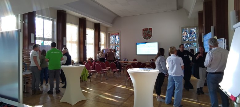In einem großen Raum mit Fenstern an einer Seite, stehen mehrere Personen in Grüppchen zusammen. Es sind Stuhlreihen aufgebaut und vorn hängt ein großer Bildschirm unter einem Gemeindewappen.