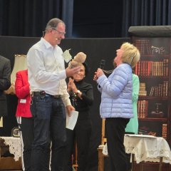 Ein Mann steht im weißem Hemd und Jeans auf einer Bühne, hält einen Teddybären in der Hand und gegenüber steht eine Frau in hellblauer Jacke und dunkler Hose mit blonden Haaren, die herzhaft lacht. Im Hintergrund sind weitere Personen und ein Bild eines Bücherregales. und ein blauer Bühnenvorhang. 