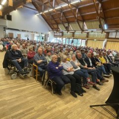 Blick in einen Saal auf ein Publikum, das auf Holzstühlen sitzt. Im Dachstuhl sieht man Holzbalken.