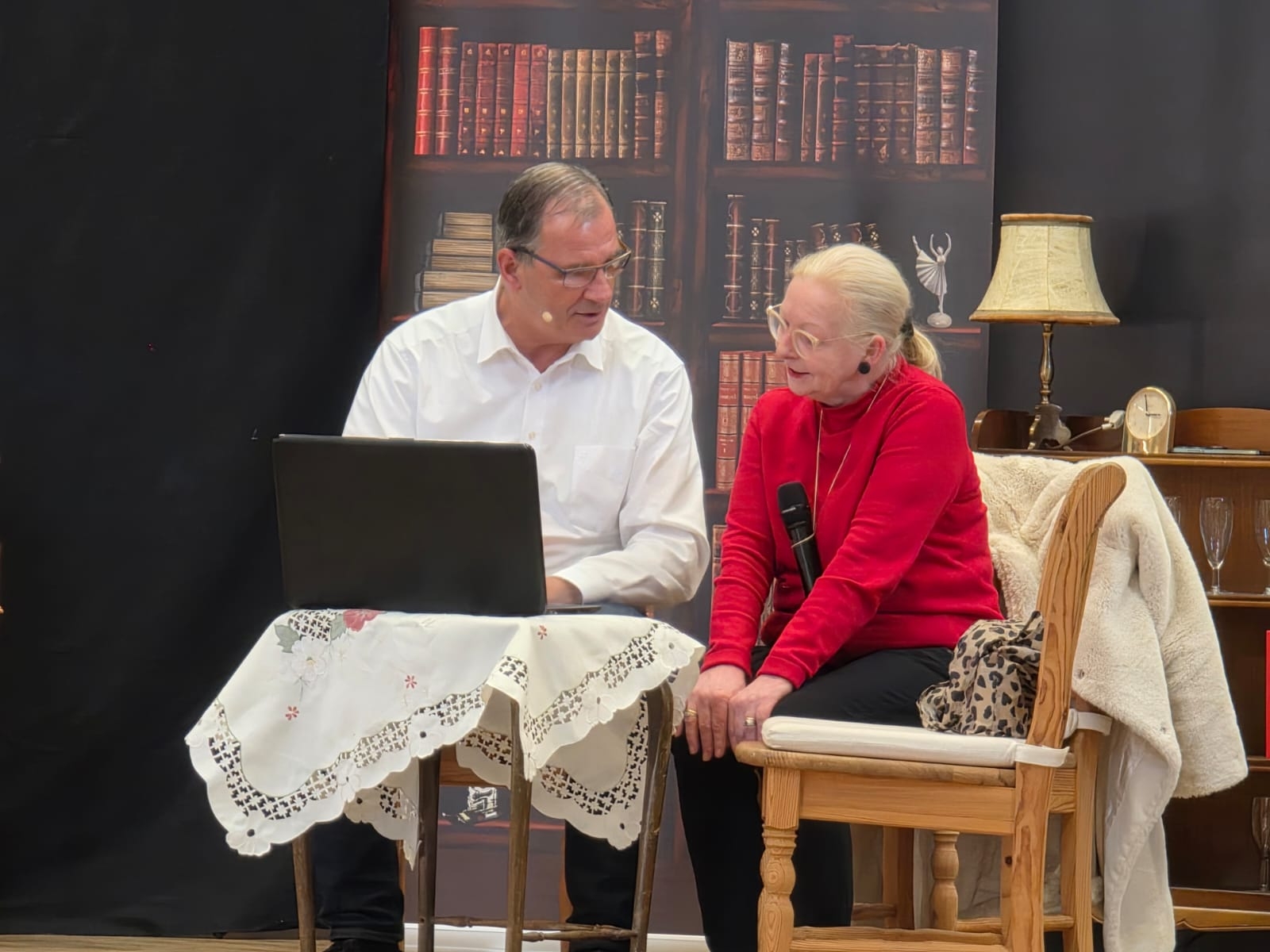Ein Mann und eine Frau sitzen auf Holzstühlen um einen kleinen Tisch herum. Auf dem Tisch ist eine weiße Tischdecke und ein aufgeklappter Laptop. Im Hintergrund ist ein Bild eines Bücherregals und ein halbhohes Regal, in dem unter anderem eine Uhr, eine Lampe und Sektgläser stehen.