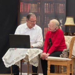 Ein Mann und eine Frau sitzen auf Holzstühlen um einen kleinen Tisch herum. Auf dem Tisch ist eine weiße Tischdecke und ein aufgeklappter Laptop. Im Hintergrund ist ein Bild eines Bücherregals und ein halbhohes Regal, in dem unter anderem eine Uhr, eine Lampe und Sektgläser stehen.