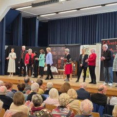 Auf einer Bühne, die im Hintergrund einen bauen Bühnenvorhang hat, stehen elf Personen vor einem Bühnenbild. Im Vordergrund sieht man das Publikum, auf Stühlen sitzend, von hinten.