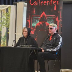 Ein Mann und eine Frau, in dunkel gekleidet, sitzen an einem Tisch mit schwarzer Tischdecke. Hinter ihnen ist ein düsteres Bild mit den roten Buchstaben "Callcenter" und eine rot-schwarze Person von hinten, die die Kaputze des Pullis aufhat und an einem PC mit großem Bildschirm sitzt, an dem viele Datne zu sehen sind.