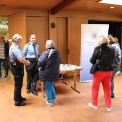 Ein Eingangsbereich mit Holzschiebetüren mit mehreren Personen und zwei Polizistinnen und einem Banner der Polizei und ein weiterer Stand mit Pult und Banner, hinter dem eine Frau steht.