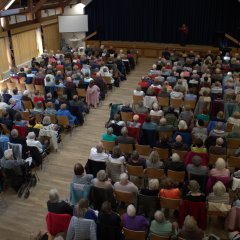 Blick von oben auf ein Publikum, das auf Holzstühlen sitzt und auf eine Bühne mit blauem Bühnenvorhang schaut. Auf der Bühne ist eine Frau mit Mikrofon.