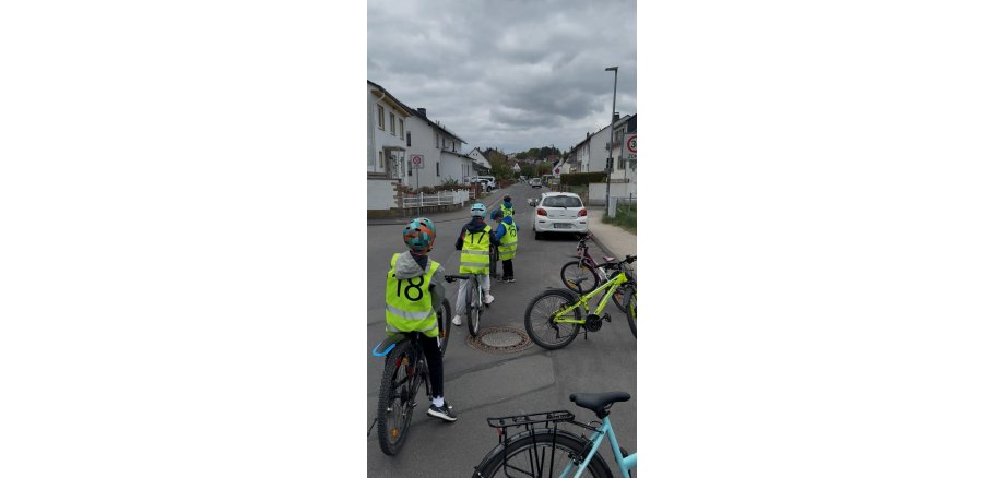 Auf einer Straße mit einer 30er-Zone im Wohngebiet parkt ein weißes Auto. Auf der Straße sieht man vier Kinder. Drei Kinder sitzen auf Ihren Fahrrädern und ein Kind steht neben seinem Fahrrad. Sie tagen Helme auf dem Kopf und gelber Warnfeste mit Zahlen auf dem Rücken. Am Straßenrand stehen drei abgestellte Fahrräder.