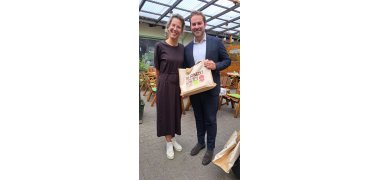 Ein Mann im blauem Anzug und weißem Hemd steht nehmen einer Frau im langemd braunem Kleid mit langen Haaren und Zopf und Brille. Er hält eine beige, rechteckige Tasche in der Hand. Beide stehen in einem Wintergarten, der mit Tischen und Stühlen bestückt ist.