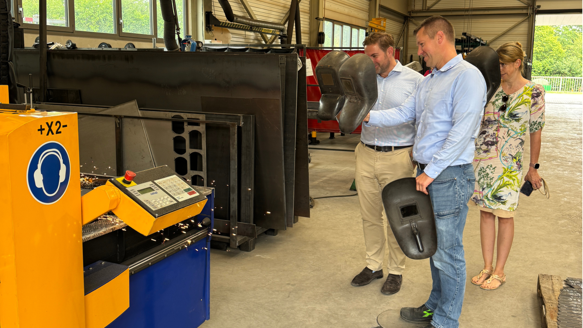 Zwei Männer und eine Frau stehen in einer Fabrikhalle und schauen auf eine blau-gelbe Maschine mit Bedienelement und Funken. Zum Schutz halten Sie schwarze Schweißer-Masken vor Ihr Gesicht.