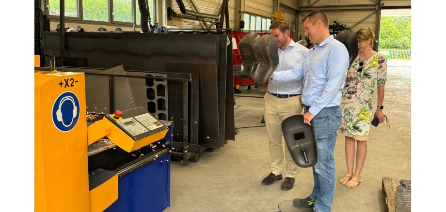 Zwei Männer und eine Frau stehen in einer Fabrikhalle und schauen auf eine blau-gelbe Maschine mit Bedienelement und Funken. Zum Schutz halten Sie schwarze Schweißer-Masken vor Ihr Gesicht.