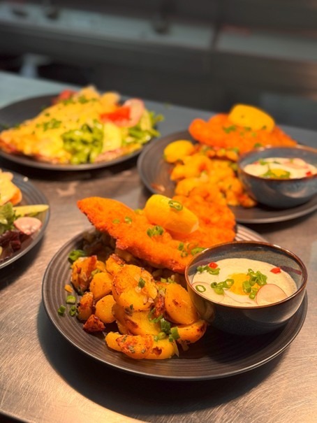 Auf einer silbernen Arbeitsplatte stehen vier runde, schwarze Teller mit Essen. Auf den rechten Tellerns sind Bratkartoffeln mit Frühlingszwiebeln und Schnitzeln und ebenso eine schwarze Schale, die mit grüner Sauce gefüllt ist und auf der eine Scheibe hart gekochtes Ei und Radieschenscheiben liegen. Auf dem Schnitzel liegt ein Viertel einer Zitrone. Links die Essen auf den Tellern sind verschwommen dargestellt.   