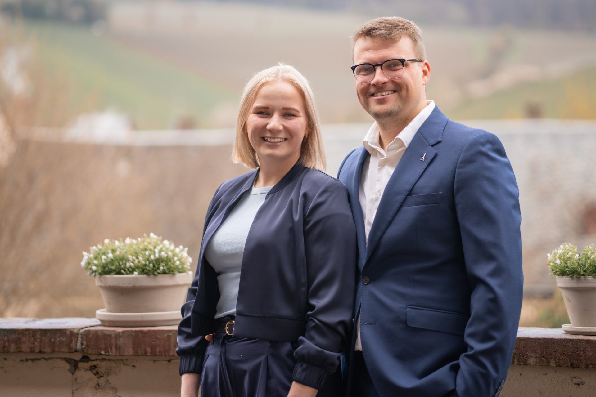 Ein junger Mann trägt einen blauen Anzug und ein weißes Hemd. Er hat eine Brille auf und hat dunkeblonde, kurze Haare. Daneben steht eine junge Frau mit blonden, schulterlangen Haaren, weißem Shirt und einer dunlkelblauen Hose und Jacke mit einem Gürtel, der eine goldene Schnalle hat. Auf der Mauer im Hintergrund stehen zwei runde Blumentöpfe mit Topfpflanzen mit weißen Blüten. Der Hintergrund ist verschwommen. 