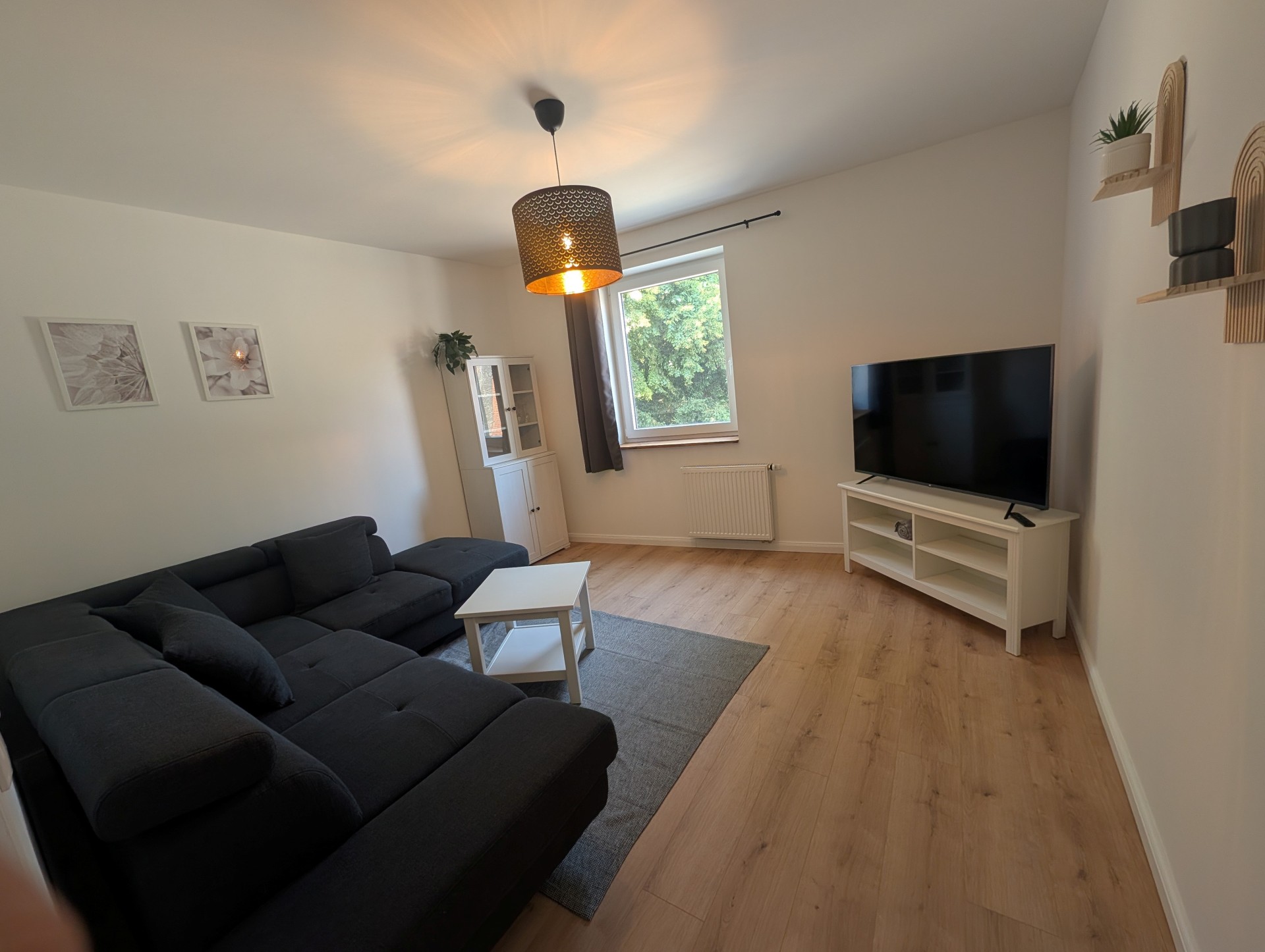 In dem Wohnzimmer steht eine L-förmige dunkelgraue Couch und ein grauer Teppich, auf dem ein weißer viereckiger Tisch steht. Ebenso steht ein weißer schrank, mit zwei Holztüren und zwei Glastüren und ein weißer TV Unterschrank, auf dem ein TV steht. Man sieht ein Fenster mit einem grauen Vorhang und zwei hellbraune Regalbretter, auf denen Blumentöpfe stehen.
