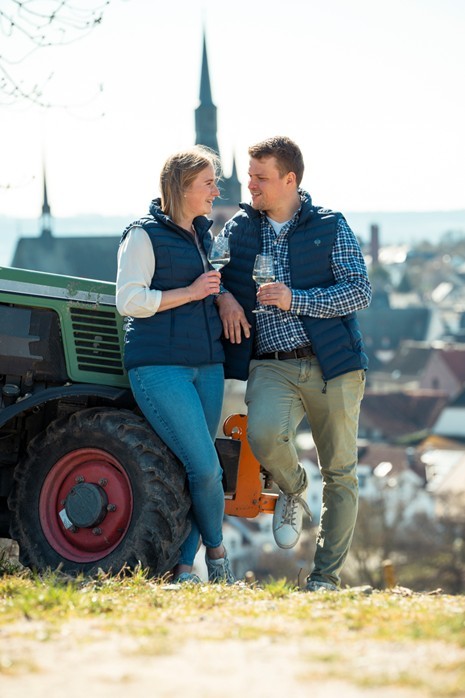 Vor einem Traktor steht eine Frau mit blonden,  schulterlagen Haaren und ein Mann mit kurzen braunen Haaren, die sich anschauen. Beide halten ein Weinglas in der Hand und tragen eine blaue Weste. Im Hintergrund ist verschwommen ein Kirchturm zu sehen. 