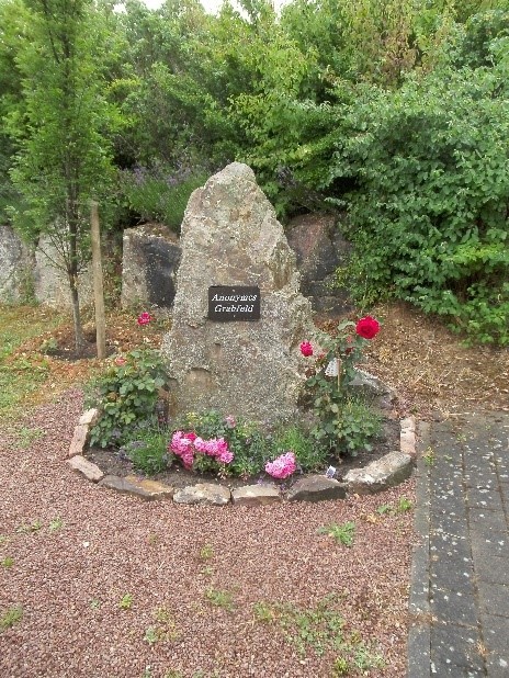An einem großen Hinkelstein ist ein dunkles, rechteckiges Schild mit weißer Schrift "Anonymes Grabfeld" befestigt. drumherum sind rot und rosa blühende Rosenund ein aus Steinen gelegter Kreis. Davor ist Kies und im Hintergrund grün belaubte Bäume.