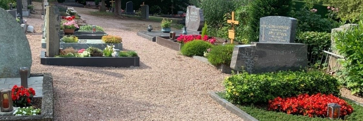 An den Kieswegen sind rechteckige mit Blumen bepflanzte Gräber mit Grabsteinen und einem Holzkreuz. Im Hintergrund Bäume, Hecken, Weinberge und ein blauer Himmel mit Wolken.