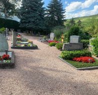 Wahlgräber An den Kieswegen sind rechteckige mit Blumen bepflanzte Gräber mit Grabsteinen und einem Holzkreuz. Im Hintergrund Bäume, Hecken, Weinberge und ein blauer Himmel mit Wolken.