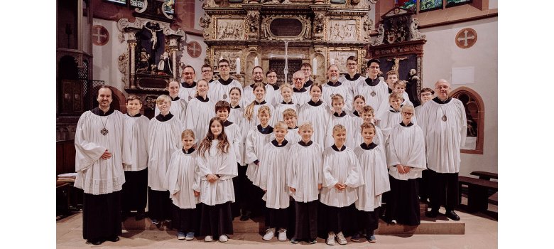 Männer der Kiedricher Chorbuben und der Kinder- und Chorschola stehen in ihren weiß- schwarzen Chorröcken vor dem Altar auf dem sechs lange, weiße Kerzen stehen.